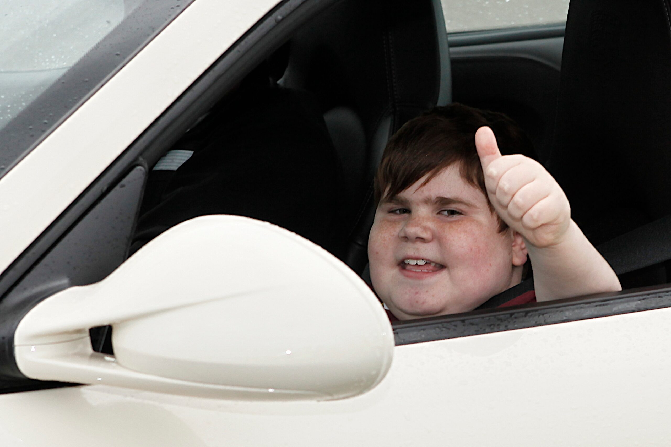 A smiling boy gives a thumbs up from the window of a white sports car